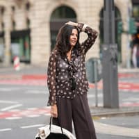 Frau mit dunkelbraunen Haaren trägt eine dunkelbraune Hose und transparente Bluse mit Muster und fässt sich mit einem Arm an den Kopf..