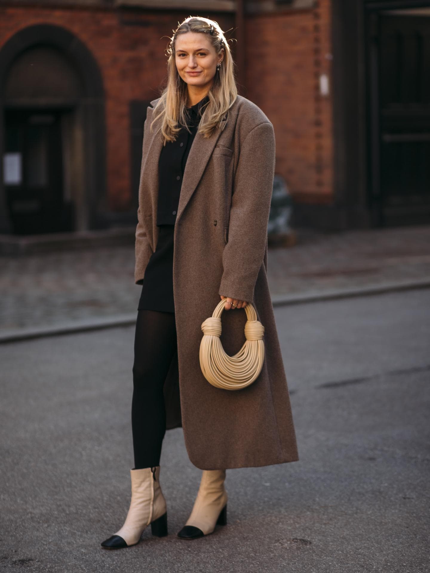 Frau trägt auf Kopenhagen Fashion Week Ankle Boots schwarzen Rock braunen Mantel und cremefarbene Tasche Ankle Boots kombinieren