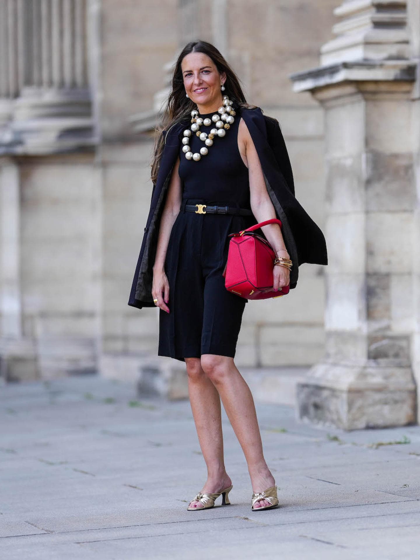 Frau mit einem schwarzen kurzen Kleid und Blazer, einer roten Tasche und Perlenkette sowie Block Heels.