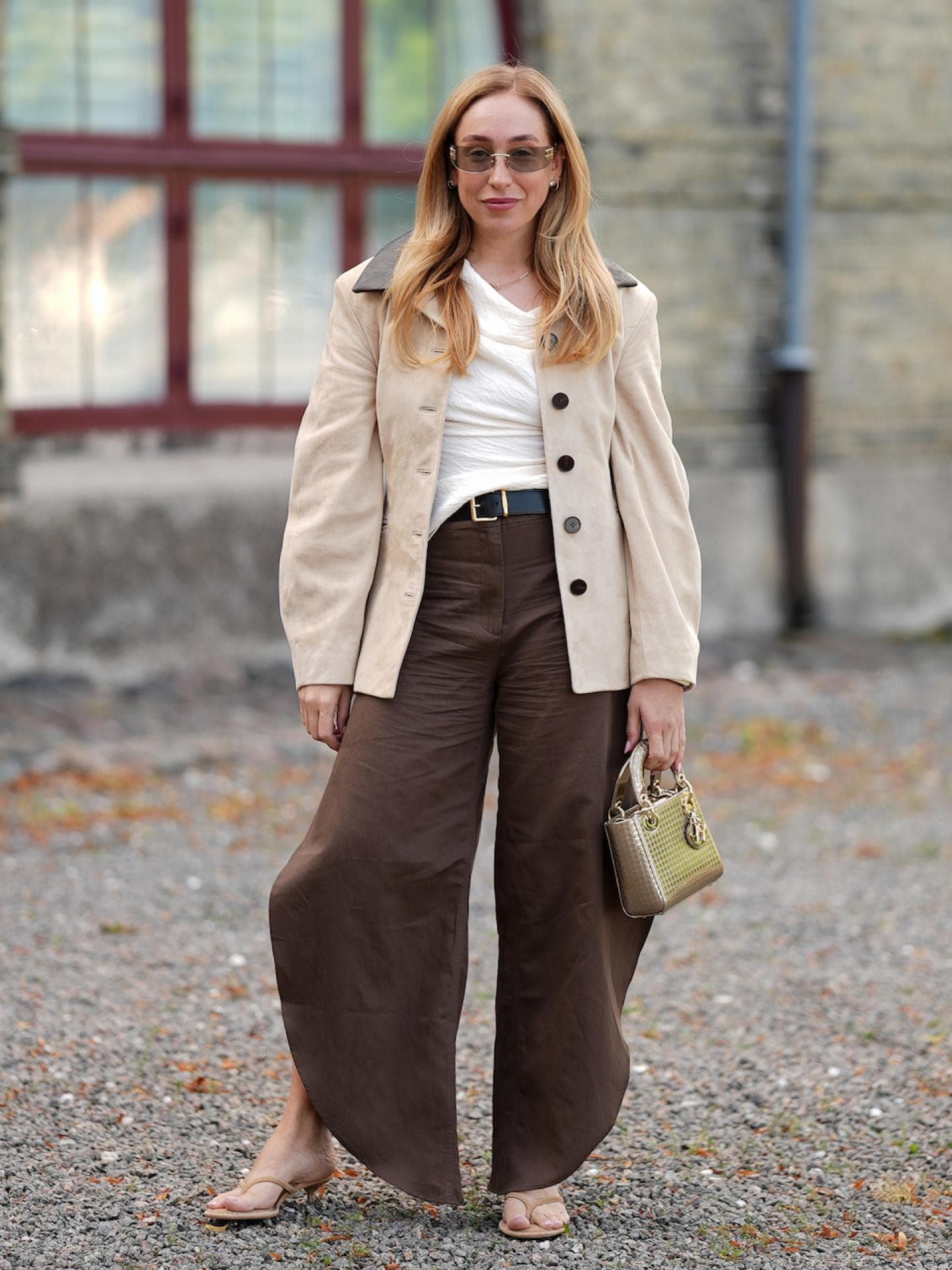 Frau mit blonden Haaren, Sonnenbrille, hellem Shirt, dunkelbrauner Hose und beigem Blazer.