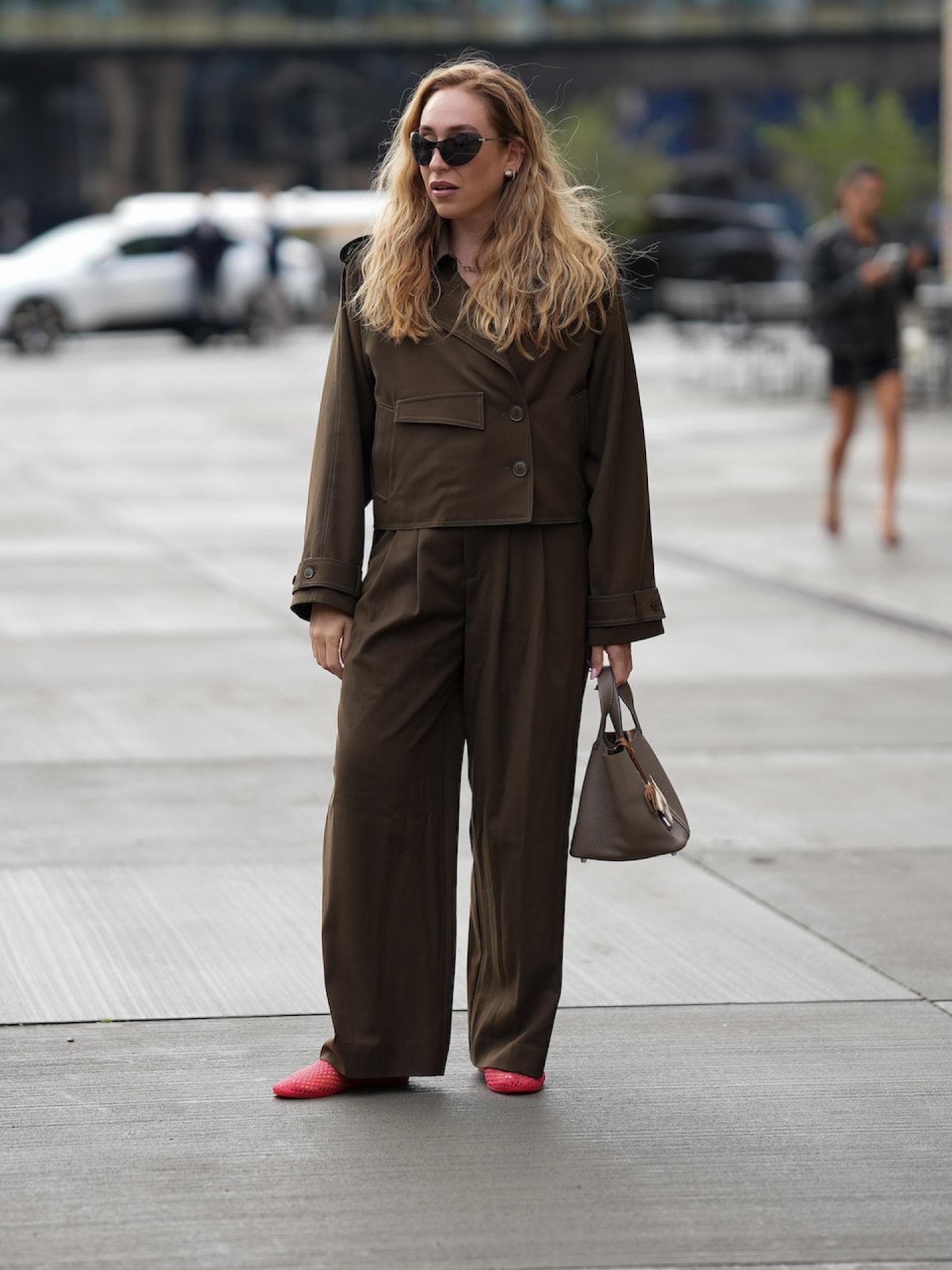 Frau mit blonden offenen Haaren und Sonnenbrille trägt eine dunkelbraune Hose und Jacke.