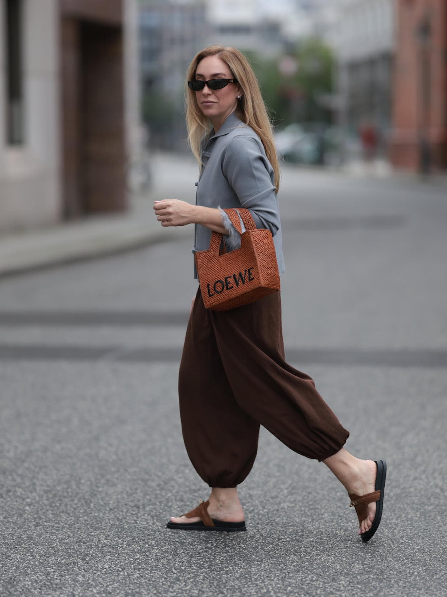 Frau mit blonden Haaren, Sonnenbrille, grauem Blazer und dunkelbrauner Hose mit FlipFlops.