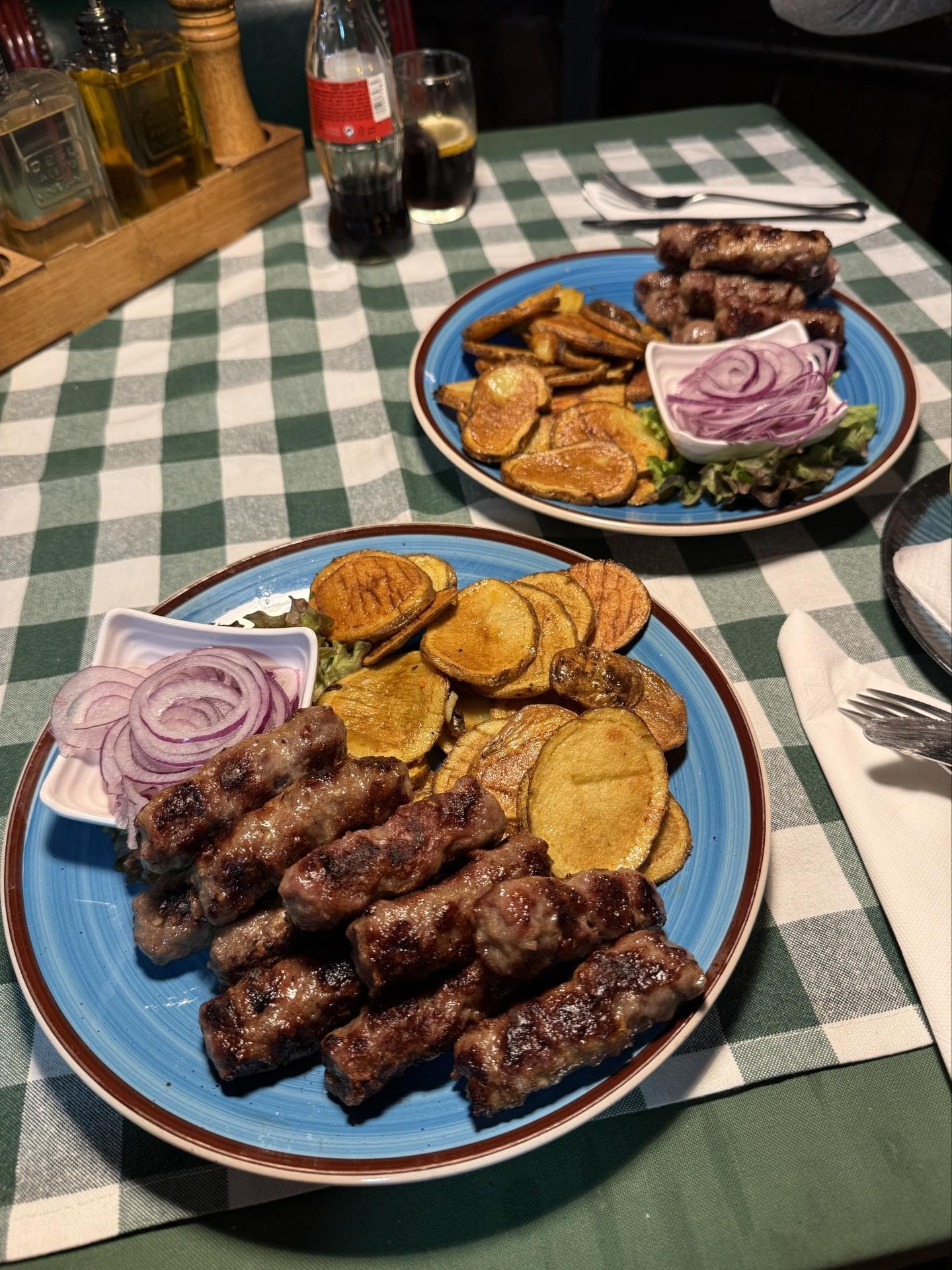 Zwei Teller auf einem Tisch mit karierter Tischdecke voll mit Ćevapi, Bratkartoffeln und Zwiebeln.