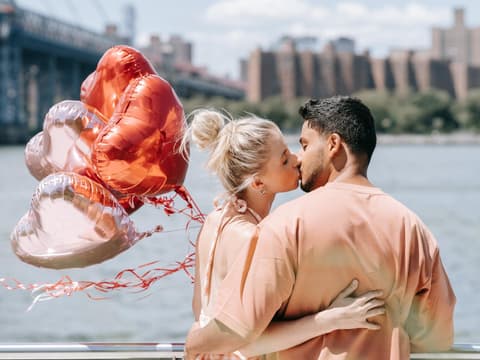 Verliebtes Paar küsst sich am Wasser und hält Herzluftballons in der Hand