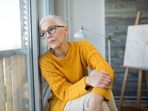 Frau mit grauen Haaren und Brille lehnt gegen das Fenster und schaut nach draußen.