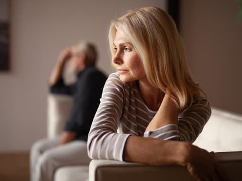 Frau sitzt auf dem Sofa und guckt zur Seite. Im Hintergrund sitzt ein Mann.