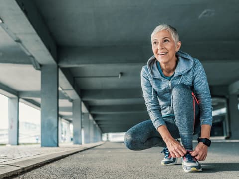 Es gibt viele Sportarten, die für einen selbst passend sein können. Eine Frau mit grauen kurzen Haaren und Sportkleidung kniet auf dem Boden und schnürt die Schuhe zu.