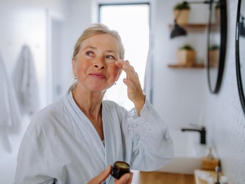 Die beste Augencreme ab 50 kann helfen, Falten zu reduzieren. Ältere Frau trägt im Bademantel eine Creme unter ihren Augen auf.