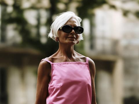 Ältere Frau mit grauen Haaren und Sonnenbrille trägt ein langes, pinkes Kleid.