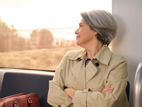 Frau mit grauen Haaren und Trenchcoat sitzt im Zug und guckt zur Seite nach draußen.