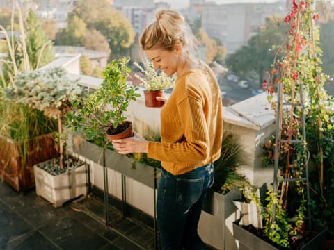 Frau steht auf einem Balkon und hält Pflanzen in der Hand.