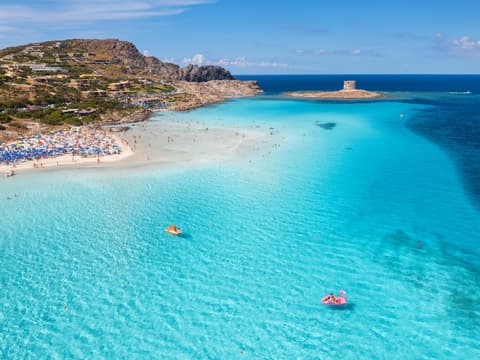La Pelosa Strand auf Sardinien mit türkisblauem Wasser, Sonnenliegen und -schirmen und Häusern im Hintergrund.