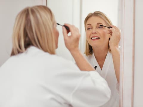 Ältere Frau mit blonden Haaren steht im Bademantel vor dem Spiegel und trägt Mascara auf.
