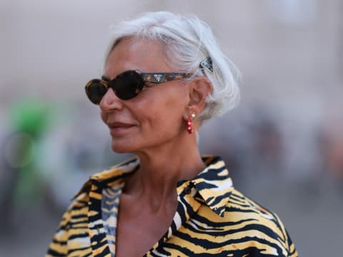Frau mit grauen kurzen Haaren, einer Sonnenbrille und einem getigerten Oberteil guckt zur Seite.