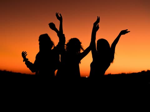 Drei Frauen tanzen ausgelassen vor dem Sonnenuntergang.
