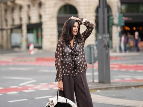 Dunkelbraune Hosen liegen im Trend. Frau mit dunkelbraunen Haaren trägt eine dunkelbraune Hose und transparente Bluse mit Muster und fässt sich mit einem Arm an den Kopf..