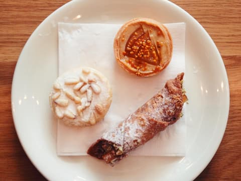 Drei italienische Backwaren (Cannoli, Torta della Nonna) auf einem weißen Teller, von oben nach unten auf einem Holztisch geschossen.