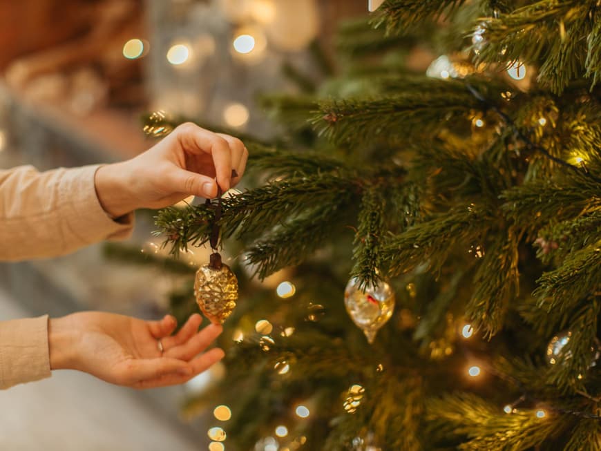 Lohnt sich ein künstlicher Weihnachtsbaum? Frau hängt goldenen Christbaumschmuck an einen Tannenbaum im Wohnzimmer