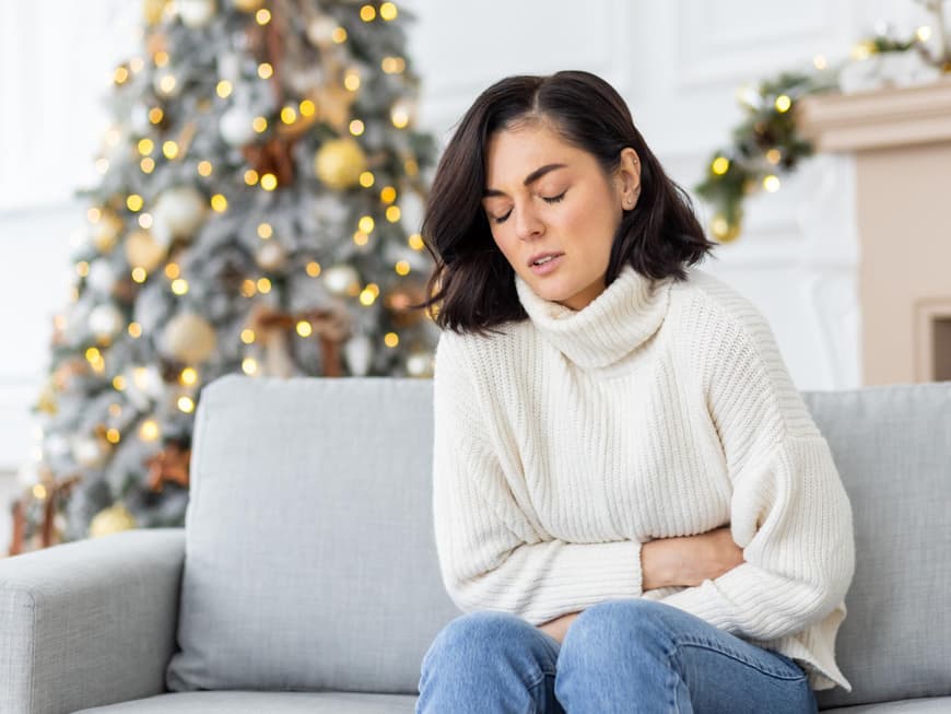 Völlegefühl in der Weihnachtszeit Frau im weißen Rollkrafenpullover sitzt auf der Couch und hat Bauchschmerzen