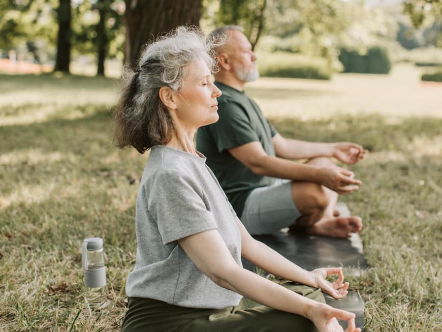 Gesunde Routine tragen zu einer höheren Lebensqualität bei. Älteres Paar macht Yoga auf einer Wiese.