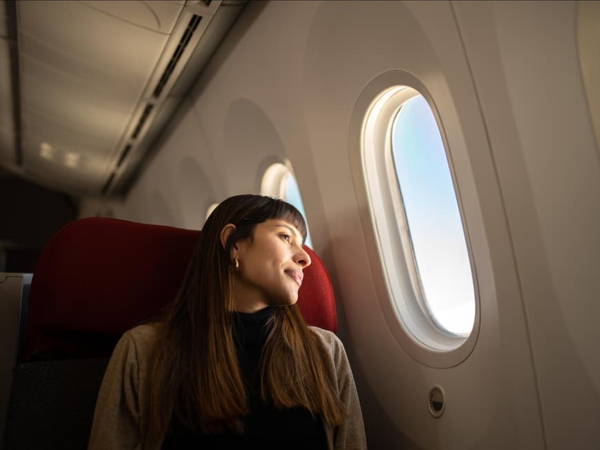 Jetlag Frau mit langen, braunen Haaren sitzt im Flugzeug und schaut aus dem Fenster