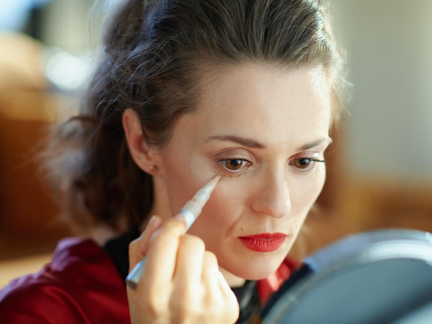 Frau sitzt vor dem Spiegel und deckt ihre Augenringe mit Concealer ab.