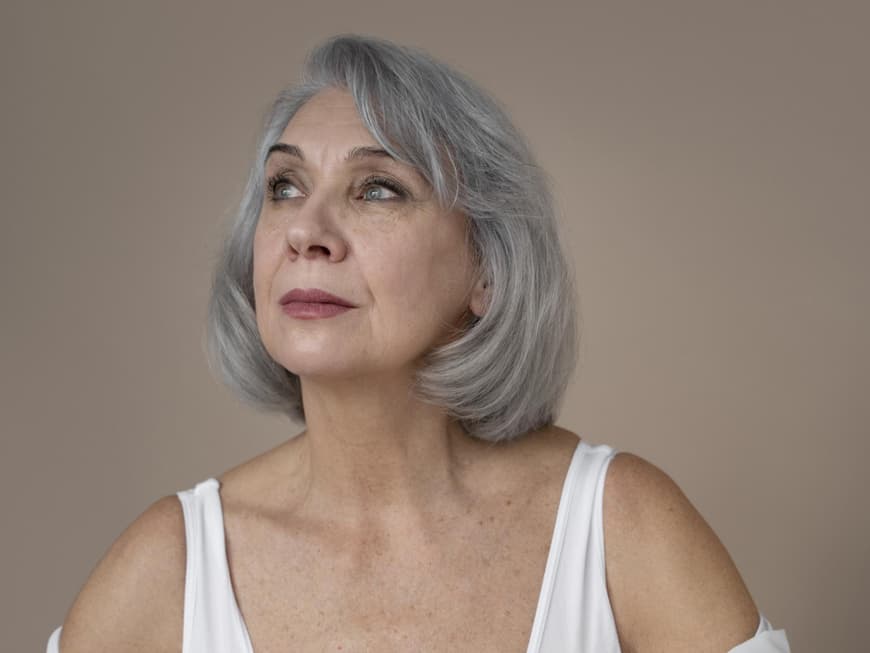 Frau mit grauen kurzen Haaren trägt eine weiße Bluse und Lippenstift.