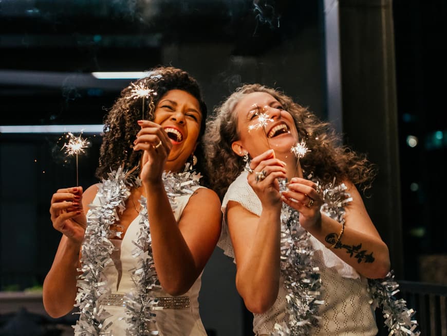 Zwei Frauen mit Wunderkerzen und Glitzerdeko um den Hals lachen.