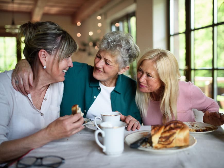 Drei Frauen sitzen am Tisch und sprechen und essen Kuchen.