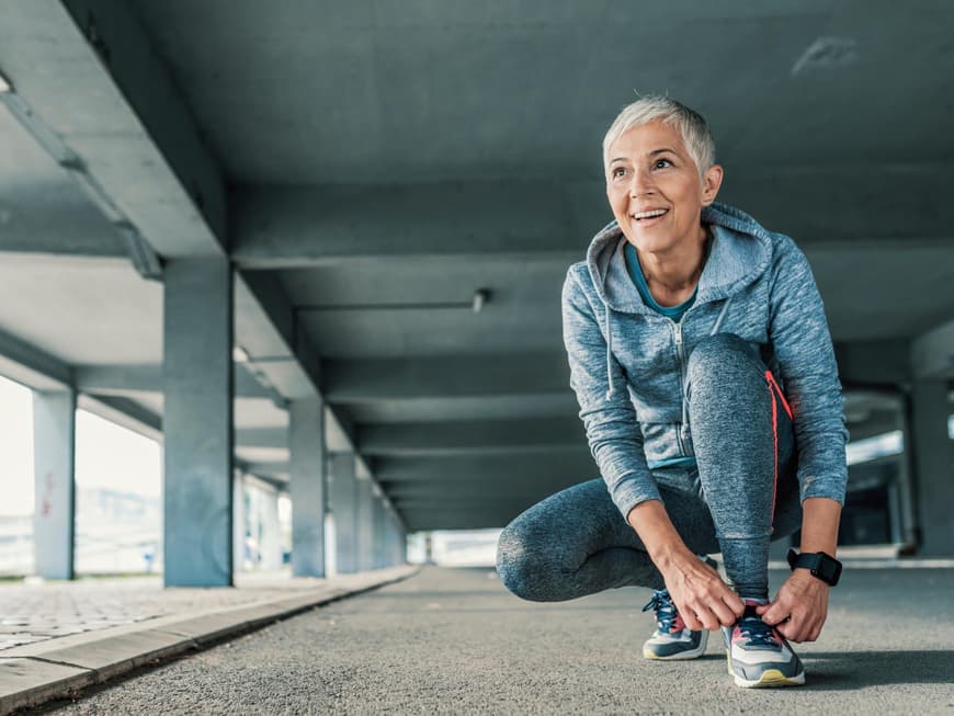 Es gibt viele Sportarten, die für einen selbst passend sein können. Eine Frau mit grauen kurzen Haaren und Sportkleidung kniet auf dem Boden und schnürt die Schuhe zu.