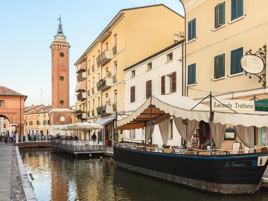 Comacchio zeichnet sich durch viele Gassen und Kanäle aus. Trattorie und Häuser mit Blick auf den Kanal, der durch Comacchio fließt. Im Hintergrund plaudern Einheimische