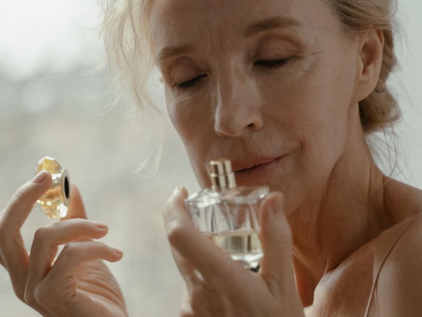 Eine ältere Frau riecht mit geschlossenen Augen an einem Parfum Flakon.