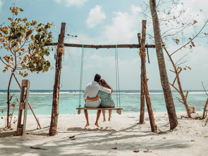 Pärchen von hinten sitzt Arm in Arm auf einer Schaukel am Strand.
