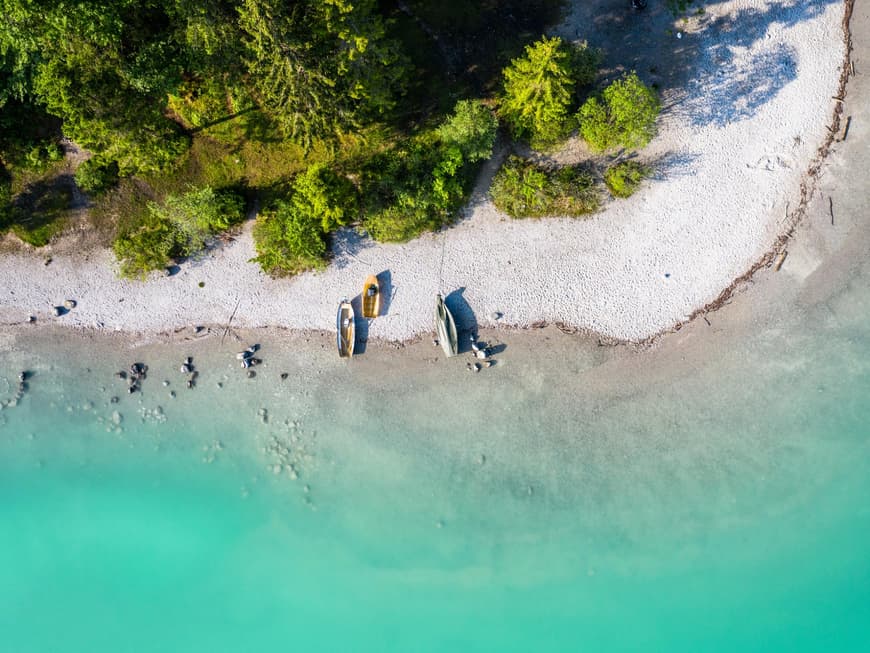 Der Strand vom Walchensee zeigt, wie schön unberührte Natur sein kann. Ein Strand mit türkisblauem Wasser, Fischerbooten und Nadelbäumen von oben.