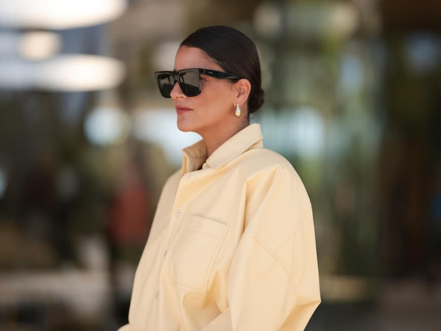Frau mit einer gelben Jeansbluse, Sonnenbrille und einem niedrigen Dutt mit kurzen Haaren.