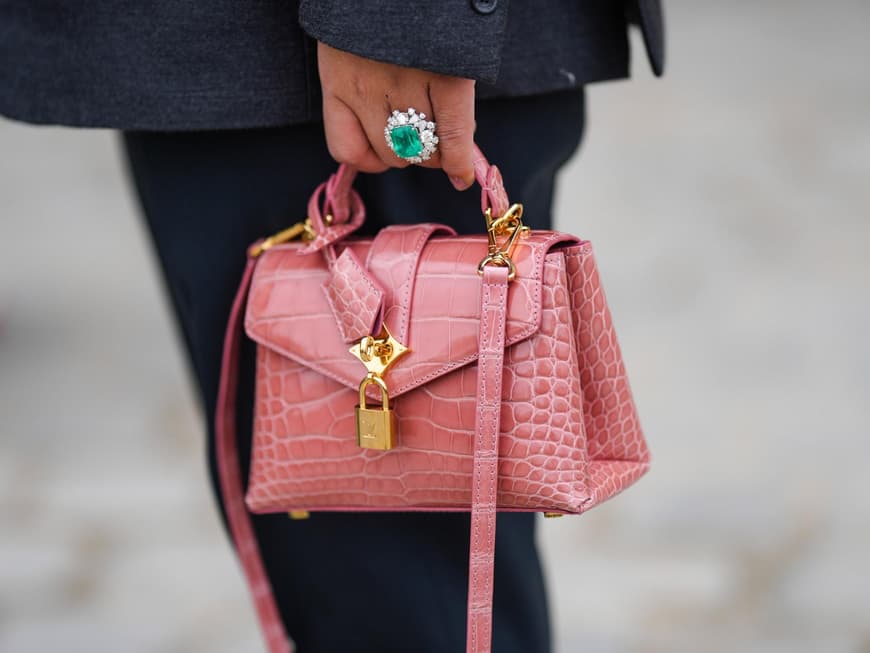Eine kleine rosane Tasche wird von einer Hand mit einem Smaragd-Ring gehalten.