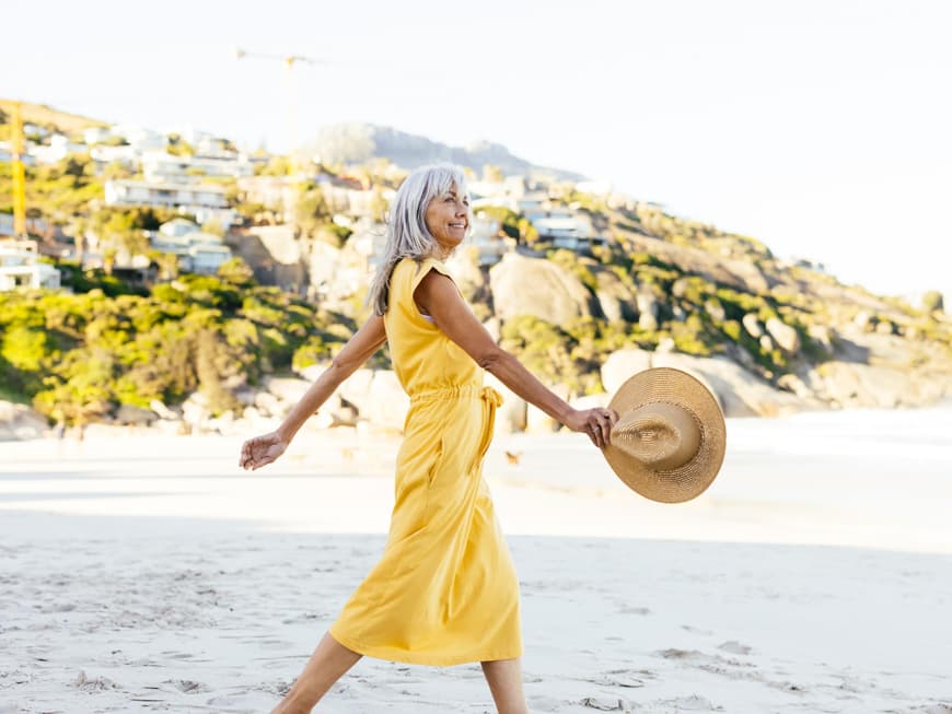 Frau mit grauen Haaren trägt ein langes, gelbes Kleid und spaziert am Strand mit einem Sonnenhut.
