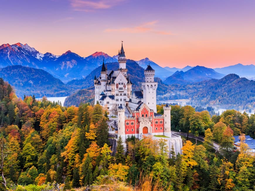 Das Schloss Neuschwanstein zwischen Bäumen mit Gebirge im Hintergrund bei Sonnenuntergang.