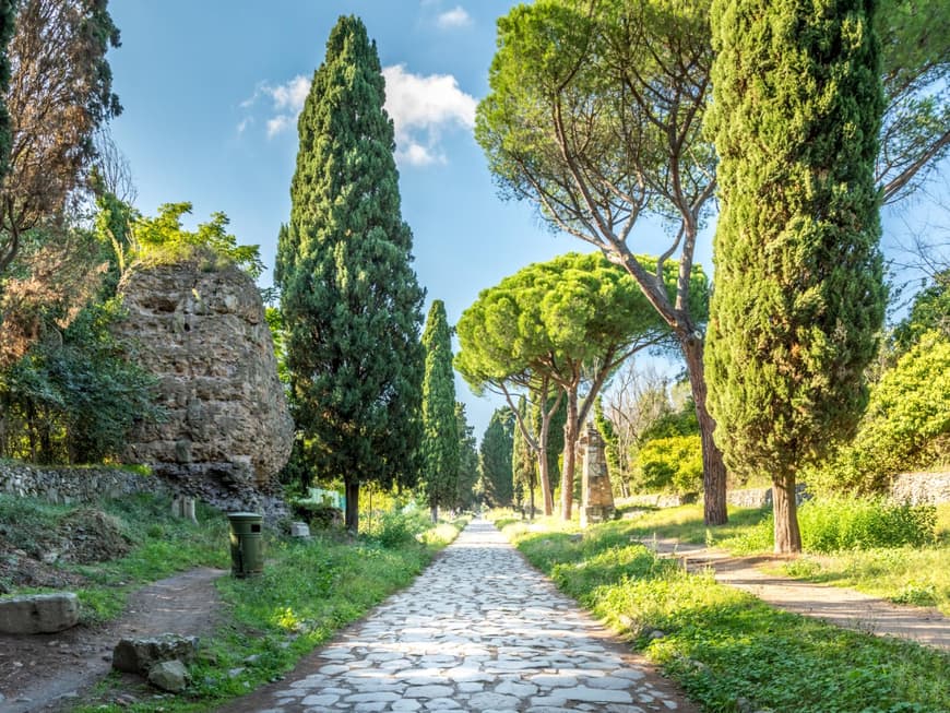 Die Via Appia Antica erstrahlt in prachtvollem Grün. Eine grüne Landschaft mit einem steinigen Weg umringt von verschiedenen Bäumen.
