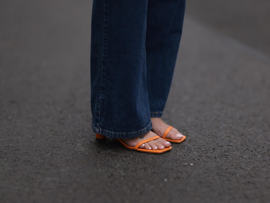 Nackte Füße in orangenen Sandalen mit hellem Nagellack und einer langen, dunklen Jeans.