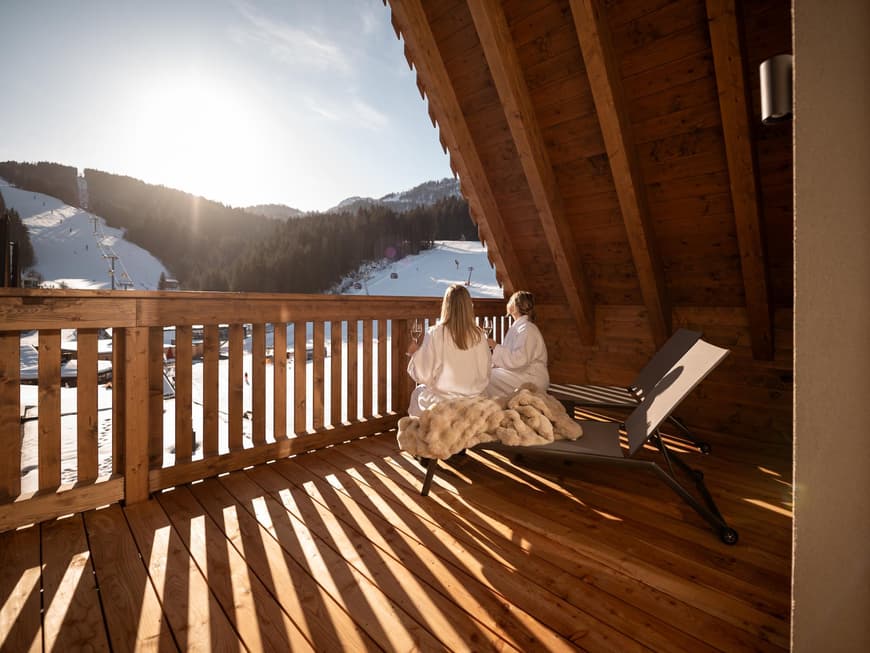 Zwei Frauen sitzen auf einem Holz-Balkon mit Bademänteln und trinken Wein. Der Ausblick ist eine Landschaft in den Bergen mit Schnee.