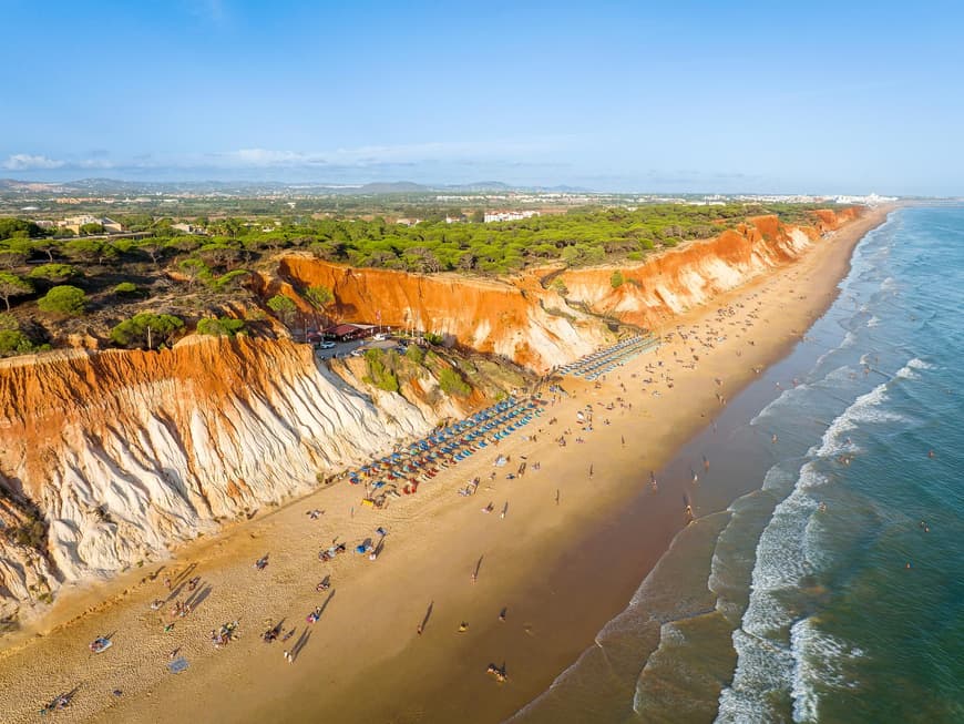 Der Praia da Falesia gilt als einer der schönsten Strände in Portugal. Ein Strand mit rötlichen Felsen, Bäumen darauf und Sonnenschirmen und -liegen am Strand.
