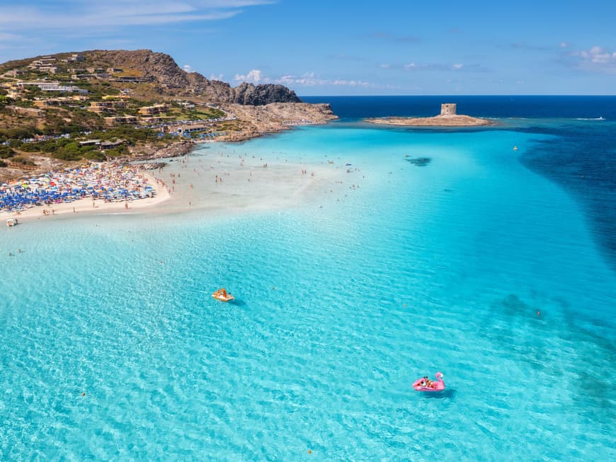 La Pelosa Strand auf Sardinien mit türkisblauem Wasser, Sonnenliegen und -schirmen und Häusern im Hintergrund.