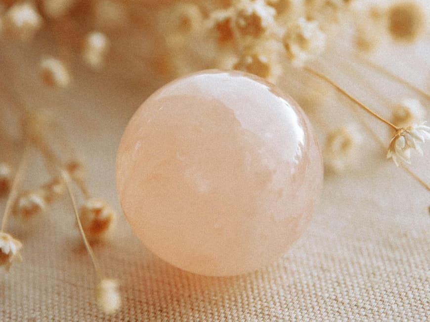 Portaltage sind im Sommer 2025 für einige wichtig. Eine kleine rosafarbene Steinkugel liegt auf einem Tisch mit beigefarbenen Trockenblumen drum herum.