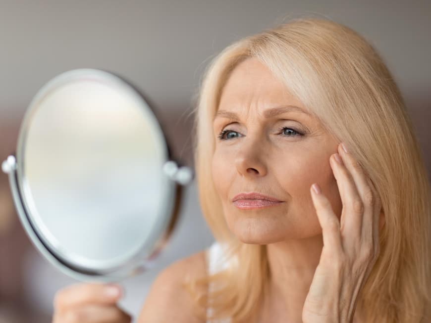 Blotting Sticks können bei glänzender Haut Wunder bewirken. Ältere Frau schaut skeptisch in einen Handspiegel und fässt sich mit der Hand ins Gesicht.