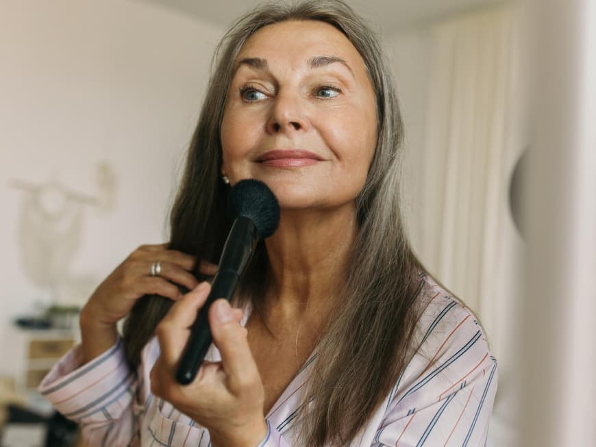 Ältere Frau mit grauen Haaren sitzt vor einem Spiegel und geht mit einem Puderpinsel über ihr Kinn.