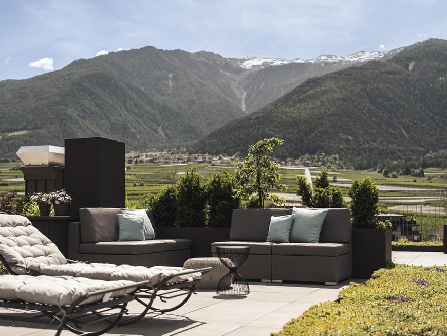 Im Matill Hotel lässt es sich ordentlich abschalten. Ein Blick auf eine Terrasse mit Liegen und Sesseln und grünen Wiesen und Bergen im Hintergrund.