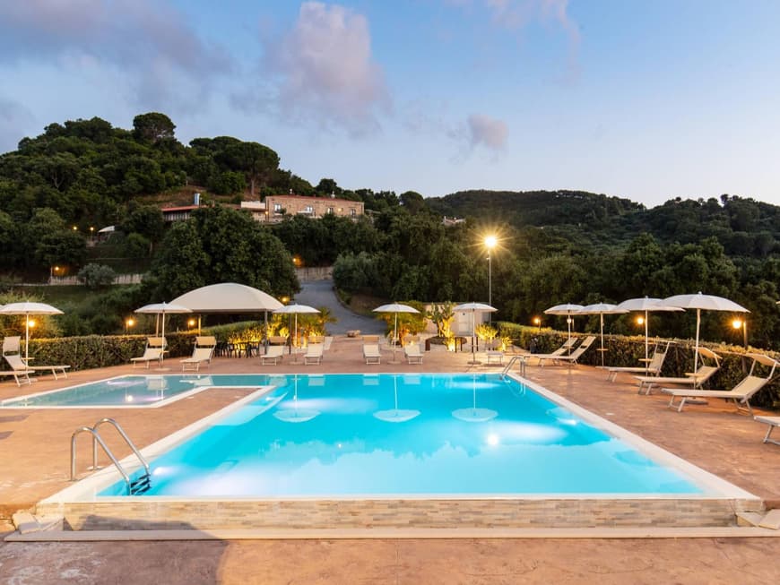 Ein Blick auf einen beleuchteten Pool mit einigen Liegen drum herum und Bäumen sowie einer Villa im Hintergrund.