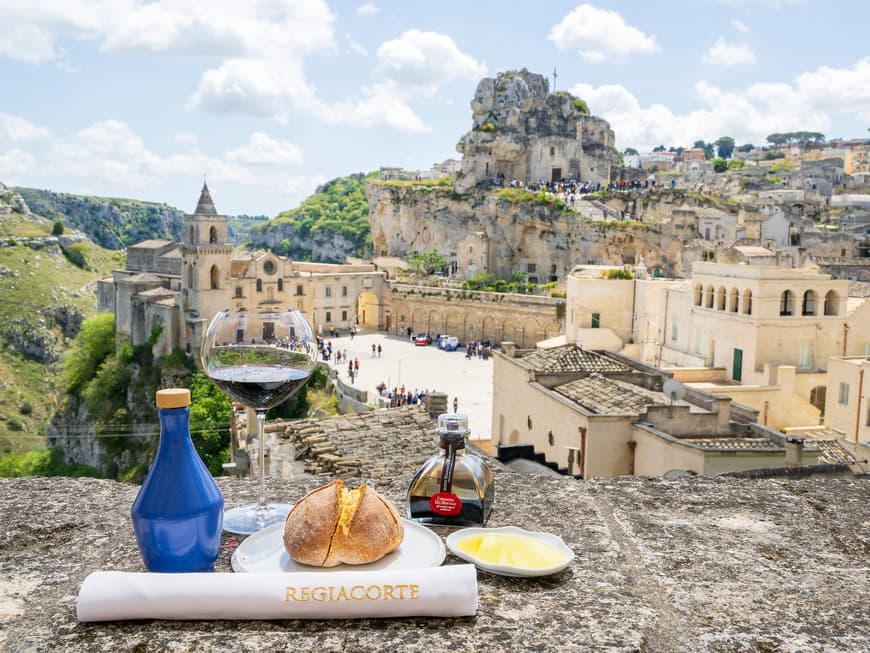 Das Restaurant Riegiacorte in Matera liegt an einer traumhaften Kulisse. Ein Brötchen und ein Weinglas auf einer Mauer mit einer Burg im Hintergrund.