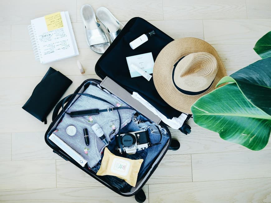 Der Koffer muss sinnvoll gepackt werden, damit du nichts vergisst. Koffer auf dem Boden mit einer Kamera, Kleidung, Beauty-Produkten, Schuhen und einem Hut.